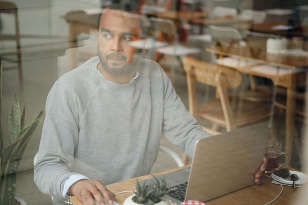 Photo of a man sitting in a cafe with a laptop on a table looking away in the distance
